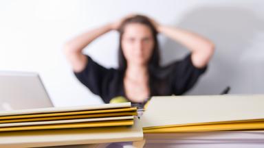 Überforderte Mitarbeiterin im Büro, die verzweifelt auf einen Stapel voll Papierkram schaut 