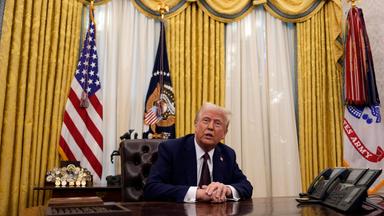 Trump sitz am Schreibtisch, Fahnen im Hintergrund