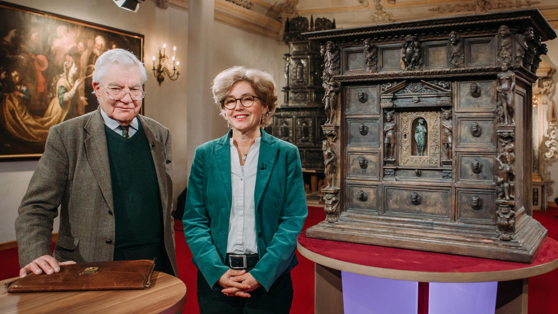 Ein Paar steht vor einem mit Figuren verzierten, antiken Holzschränkchen.