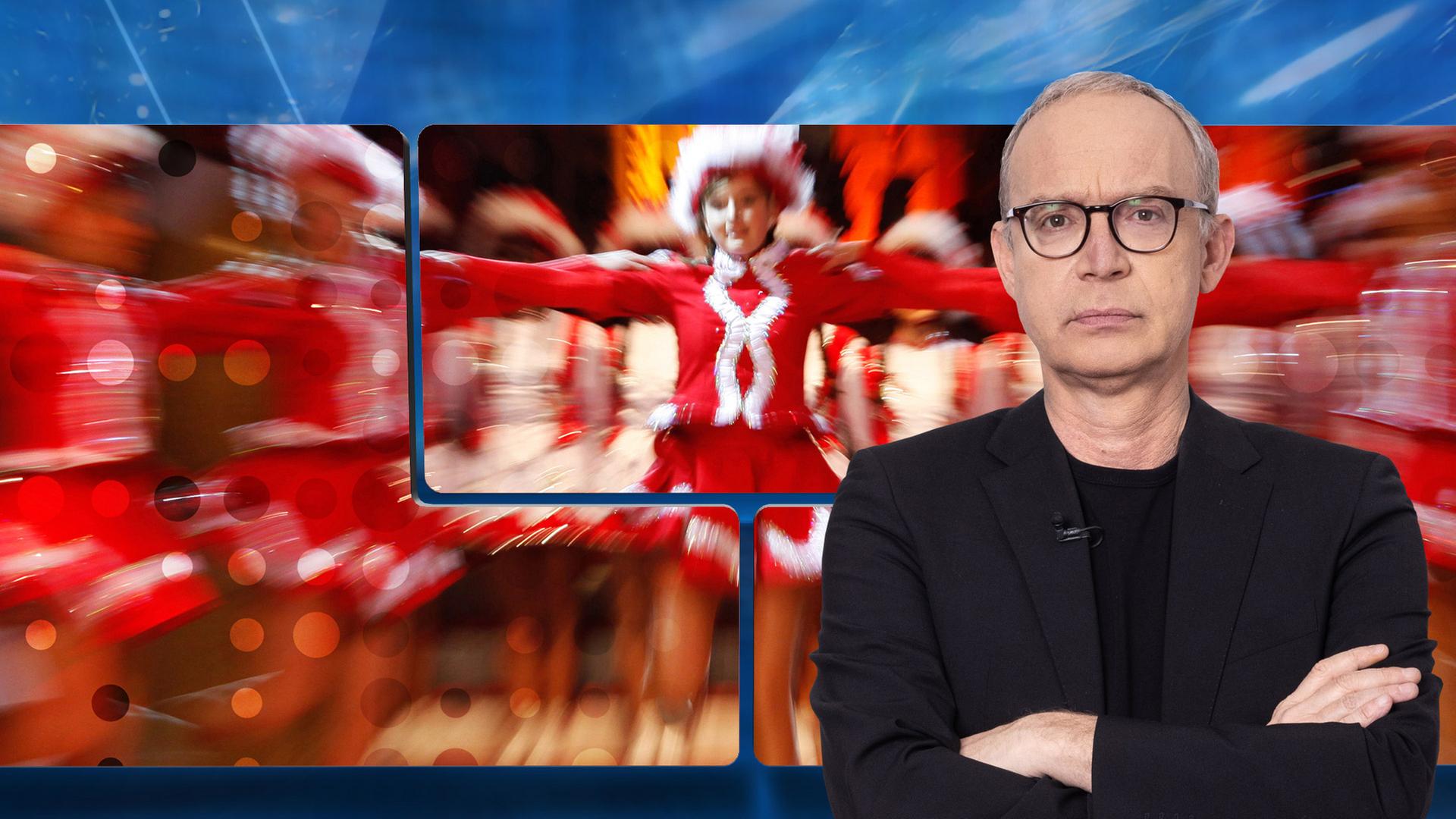 Der Moderator mit strafendem Blick vor einer Videowand, auf der ein unscharfes Bild von tanzenden Funkenmariechen zu sehen ist.