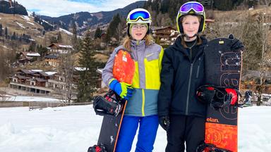 Zwei junge Mädchen stehen mit Snowboard im Schnee.