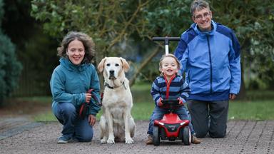 Ulrike und Christian Karges knien im Freien neben ihrem lachenden Sohn Janis, der auf einem roten Kinderfahrzeug sitzt. Zwischen ihnen sitzt ein heller Labrador. Alle blicken in die Kamera.