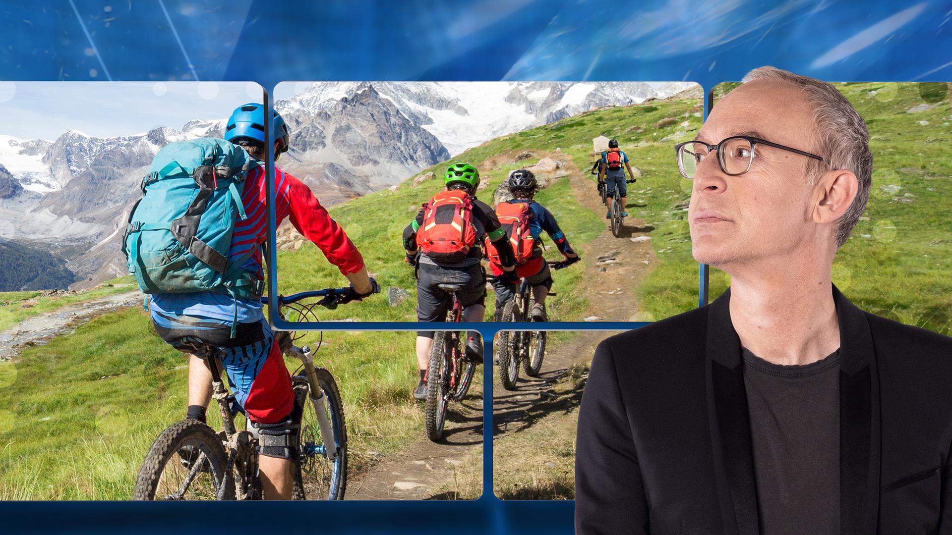 Der Moderator vor einer Videowand, auf der vier Mountain-Biker in einer Gebirgslandschaft zu sehen sind.