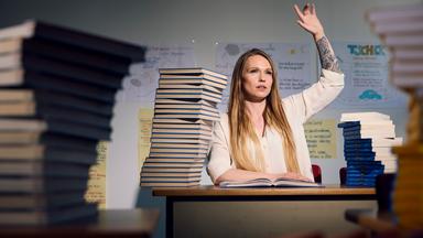 Jeder Tag ein Kampf": Sarah sitzt auf einer typischen Schulbank. Vor ihr liegt ein Berg an Schulbüchern. Sie meldet sich, augenscheinlich um eine Antwort zu geben. Im Hintergrund sind Plakate mit Lerninhalten zu erkennen.