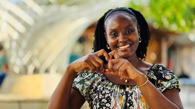 Eine Frau formt mit den Fingern ein Herz in Richtung Kamera.