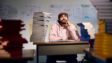 Ein Mann sitzt am Schreibtisch, links und rechts Bücherstapel. 