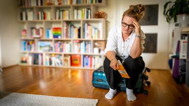 "37°Leben: Wenn die Panik mitfliegt - Flugangst besiegen": Sophie (33) sitzt auf einem Koffer in ihrem Wohnzimmer und hat ein Flugticket in der Hand. Ihr Kopf ist mit einer Hand aufgestützt und ihr Gesichtsausdruck zeigt ihre Sorge und Angst.
