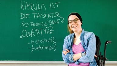 Eine Frau mit Brille und Stirnband sitzt im Rollstuhl vor einer grünen Tafel, auf der mit Kreide eine Frage zur QWERTY-Tastatur geschrieben steht. Sie lächelt in die Kamera.