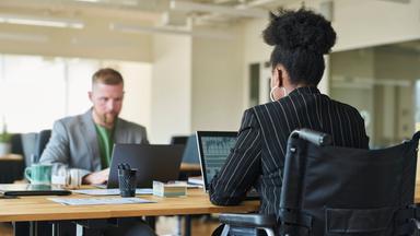 Ein weißer Mann und eine schwarze Kollegin im Rollstuhl sitzen sich an einem Schreibtisch im Büro gegenüber.