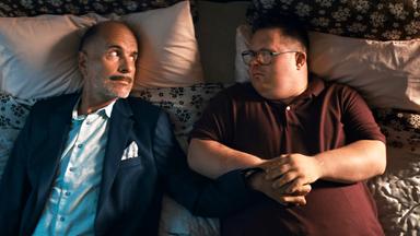 Zwei Männer liegen in Straßenkleidung auf einem Bett und schauen sich an. 