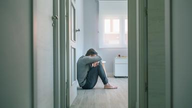 Mann sitzt an die Wand gelehnt auf dem Boden und hält sich die Hände vors Gesicht. 
