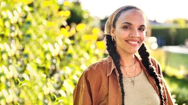 Eine Frau mit langen Zöpfen steht lächelnd vor Weinstöcken.