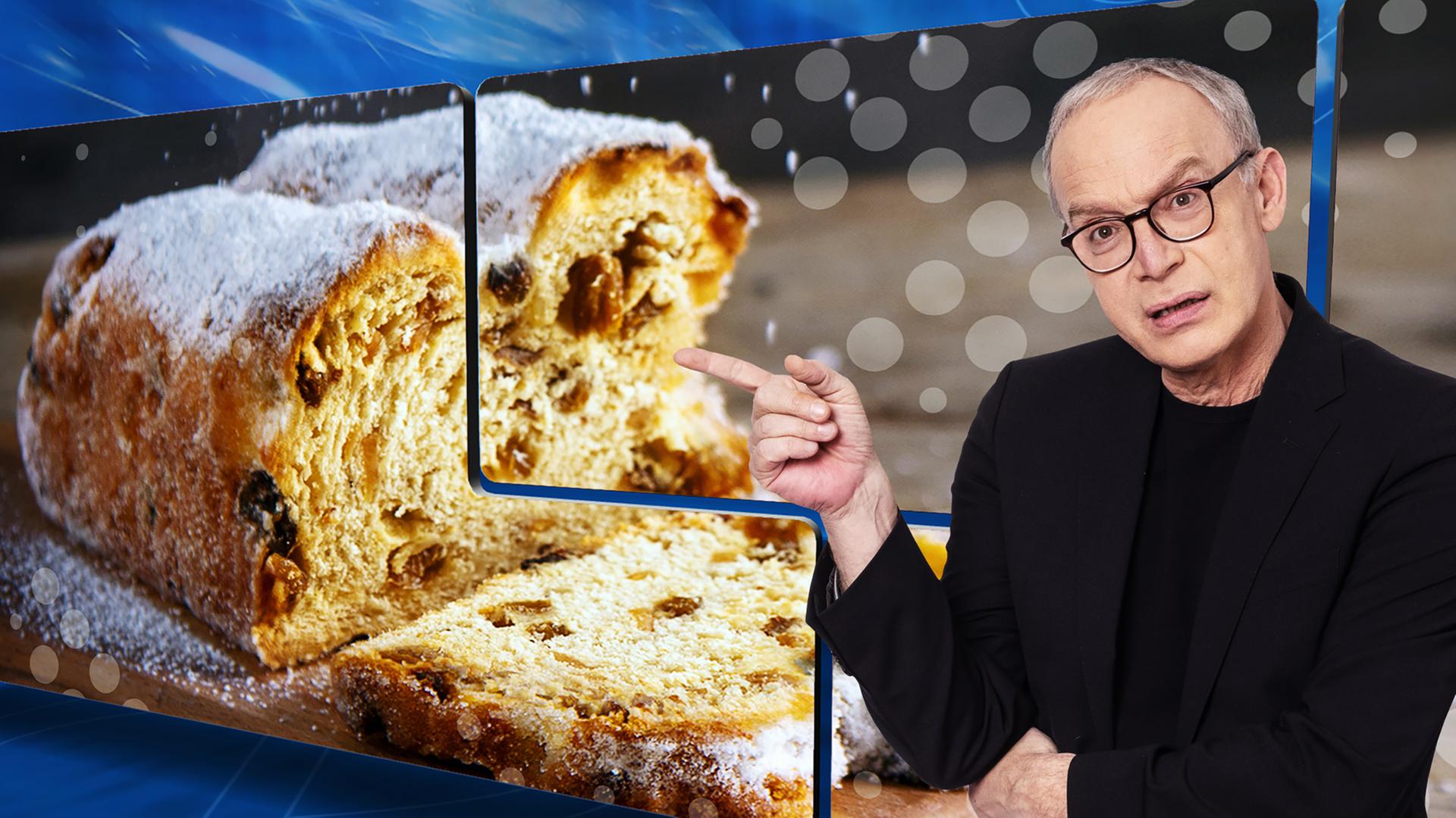 Der Moderator Christoph Süß zeigt mit dem Finger auf einen großen Weihnachtsstollen, der auf der Videowand hinter ihm zu sehen ist.