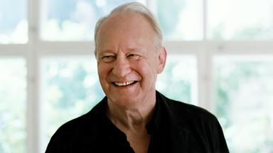 Ein lachender Mann mit weißem Haar vor einem Sprossenfenster.