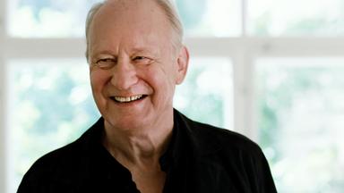 Ein lachender Mann mit weißem Haar vor einem Sprossenfenster.