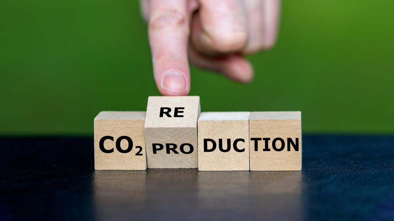Holzblöcke zeigen das Wort „CO₂ PRODUCTION“, während eine Hand einen Block mit „RE“ bewegt, sodass daraus „CO₂ REDUCTION“ werden kann.