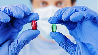 Ein medizinisch gekleideter Mann mit Mundschutz hält jeweils eine Pille in jeder Hand vor sein Gesicht