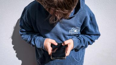 Suchtgefahr bei Jugendlichen: Politik fordert Altersbeschränkungen für Social Media, doch Experten warnen vor einem zu schnellen Verbot. (Bild zeigt eine nach unten aufs Handy schauenden Jungen im blauen Hoodie)
