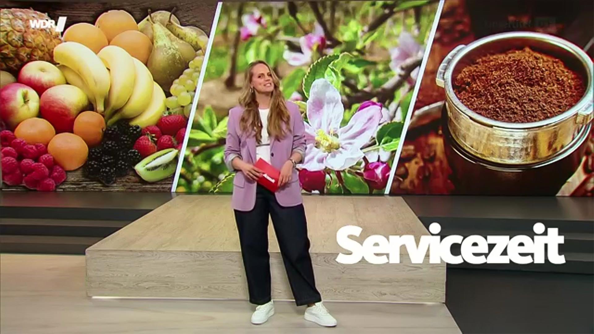 Eine junge Frau steht im Studio vor einem Hintergrundbild mit Obst, Frühlingsblumen und einem Siebträger mit Kaffeepulver