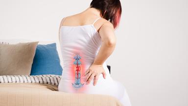 Rückenschmerzen oder Bandscheibenvorfall: Notfall oder reicht Bewegung und Therapie? (Bild: Frau sitzt auf dem Bett, hält den Rücken vor Schmerz - mit hervorgehobenem Skelett)