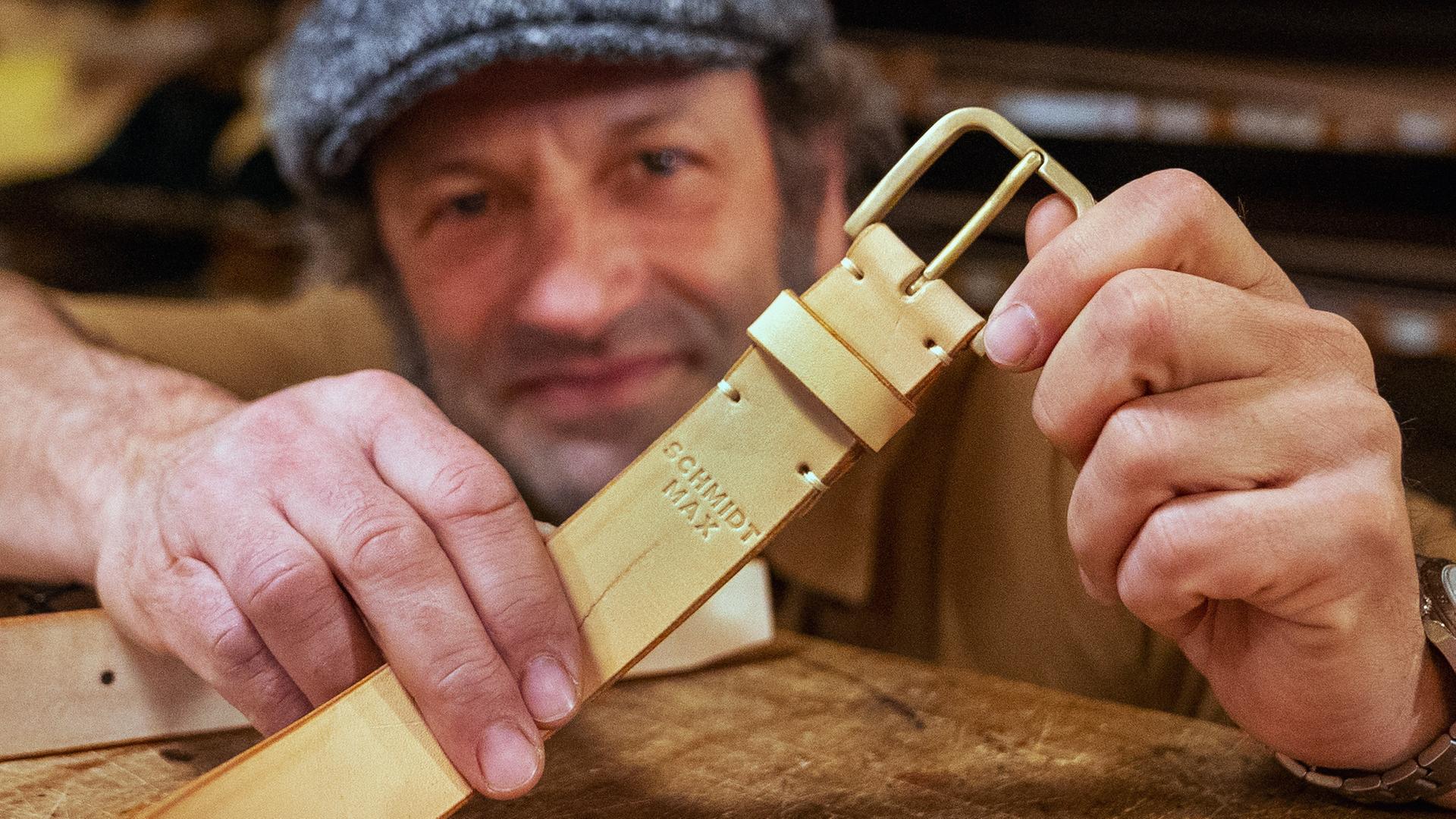 Ein Mann blickt in die Kamera und hält einen Ledergürtel, auf dem sein Name eingeprägt ist, in den Händen.