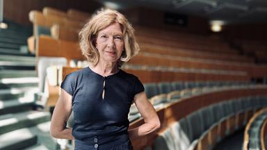 Eine Frau steht in grauschwarzer Kleidung in einem sonnendurchfluteten Hörsaal.