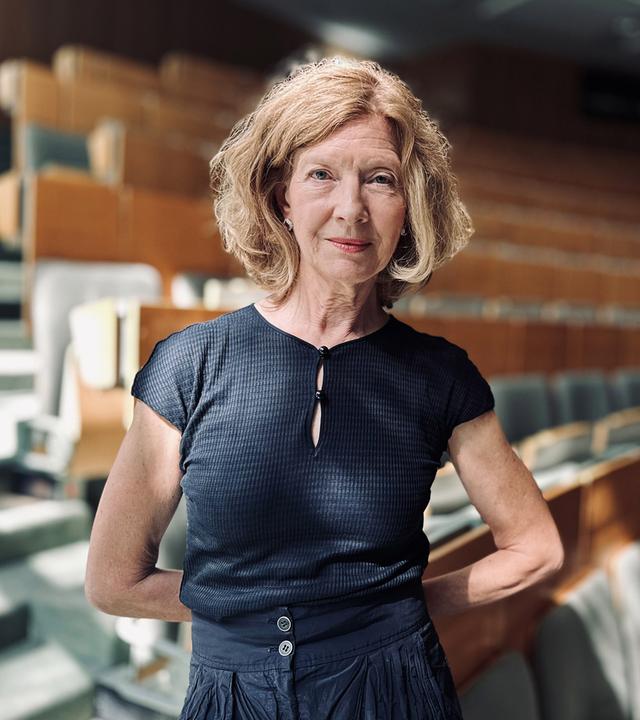 Eine Frau steht in grauschwarzer Kleidung in einem sonnendurchfluteten Hörsaal.