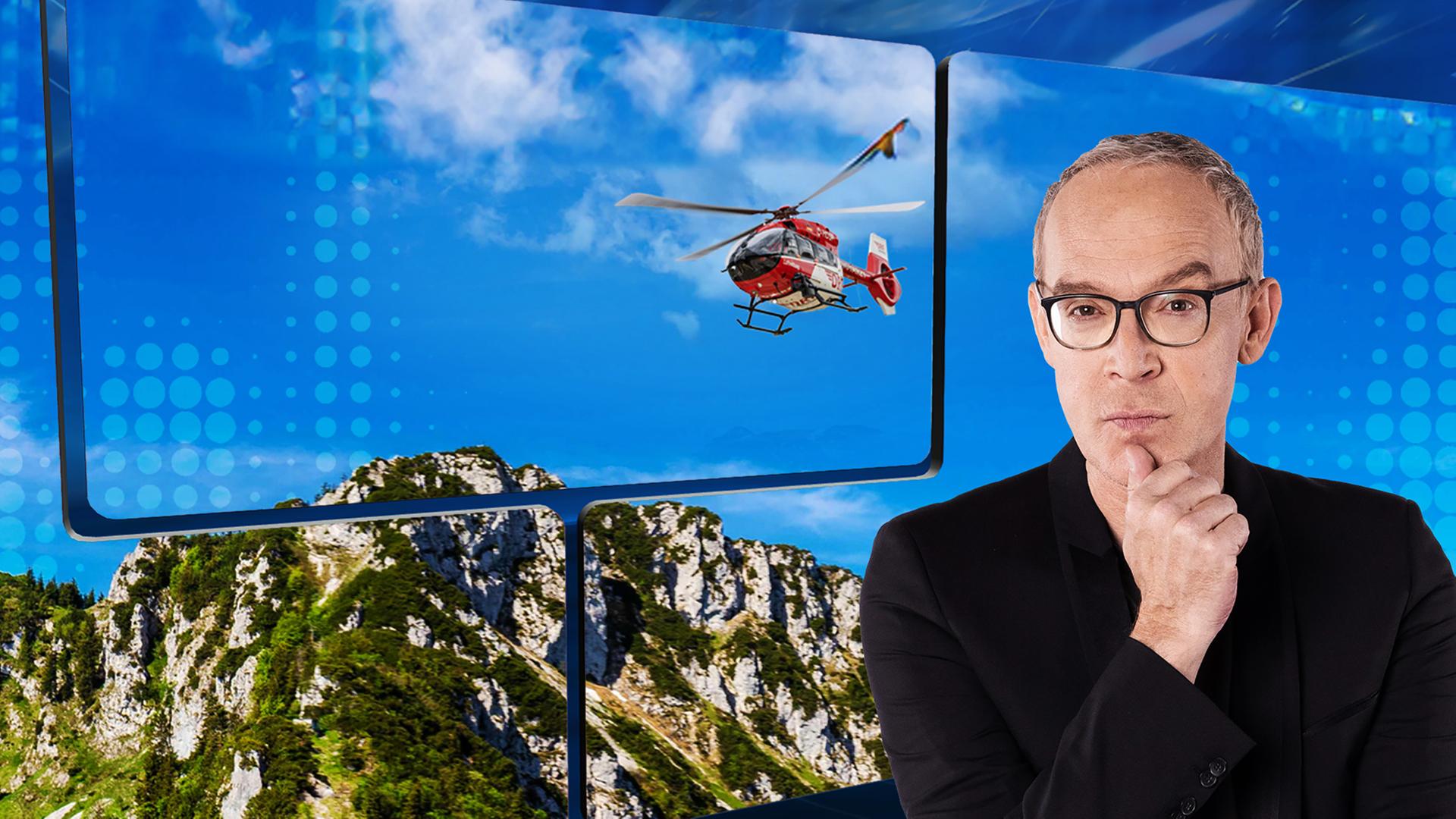 Der Moderator vor einer Videowand, auf der ein Rettungshubschrauber über einem felsigen Berg zu sehen ist.