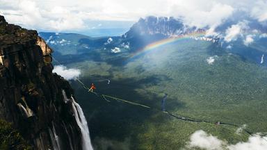 Szene aus Six Days to the Edge. Eine Person läuft auf einem Seil zwischen zwei Bergen gespannt in großer Höhe. Im Hintergrund sieht man ein Tal und ein Regenbogen.