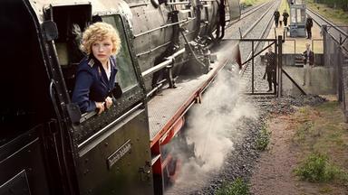Eine blonde Frau streckt den Kopf aus einer Dampflokomotive
