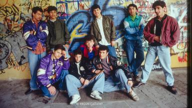 Foto einer Gruppe männlicher Jugendlicher in 1980er Jahre Klamotten vor einer mit Graffitti besprühten gelb gekachelten Wand.