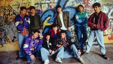 Foto einer Gruppe männlicher Jugendlicher in 1980er Jahre Klamotten vor einer mit Graffitti besprühten gelb gekachelten Wand.