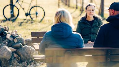 Elisa steht an ein Ortsschild aus Holz gelehnt und lächelt in die Kamera. Auf dem Schild steht "Willkommen in Quarnebeck" neben einem Wappen.
