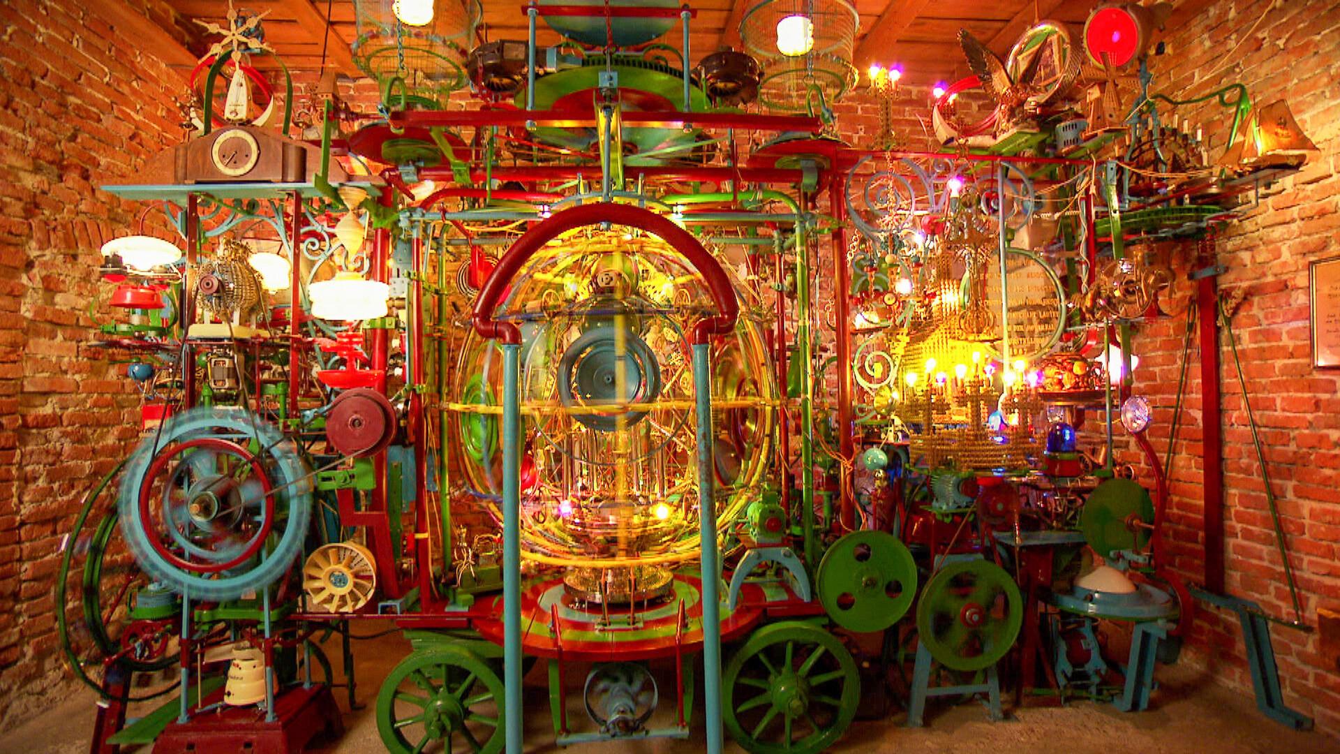 Eine komplexe, farbenfrohe Maschine aus Zahnrädern, Lampen, Röhren und Alltagsgegenständen steht in einem Raum mit Ziegelwänden; sie leuchtet, blinkt und bewegt sich.