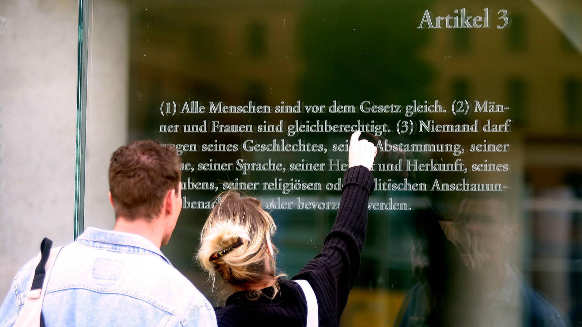 Ein Mann und eine Frau stehen vor einer Glaswand, auf der Artikel 3 des Grundgesetzes zu lesen ist.