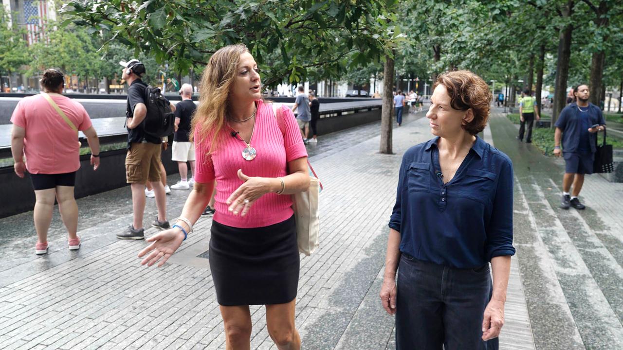 Zwei Frauen stehen im Vordergrund und unterhalten sich. Die eine trägt ein rosa Shirt, die andere ein blaues Hemd. Im Hintergrund sind Menschen, Bäume und eine Gedenkstätte sichtbar.