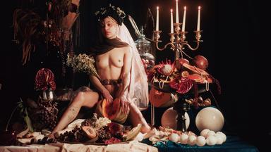 Eine nackte junge Frau mit Blumenschmuck im Haar sitzt auf einem opulent arrangierten Tisch voller Früchte und Blumen.