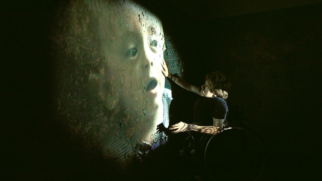 In einem abgedunkelten, hallenartigen Raum sitzt ein junger, kleinwüchsiger Mann in seinem Rollstuhl vor einer Wand, auf die groß ein Kindheitsfoto von ihm projiziert wird. Von der Wand blättert Putz ab.