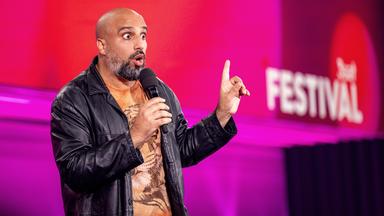 Abdelkarim steht in der Mitte des Bildes. In der rechten Hand hat er ein Mikrofon. Die linke Hand zeigt mit dem Zeigefinger nach oben. Der Hintergrund ist rot und lila.