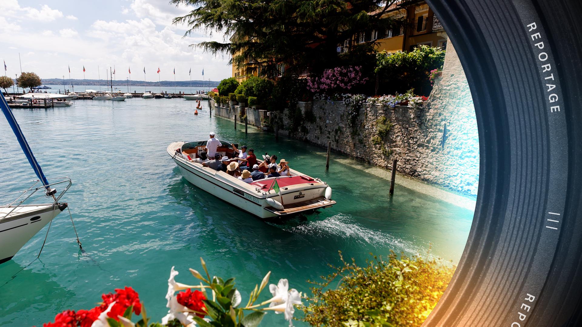 Ein Touristenboot, ein Wassertaxi, fährt an einer Mauer entlang auf dem Gardasee. Im Vordergrund blühen Blumen, im Hintergrund liegen weitere Schiffe.