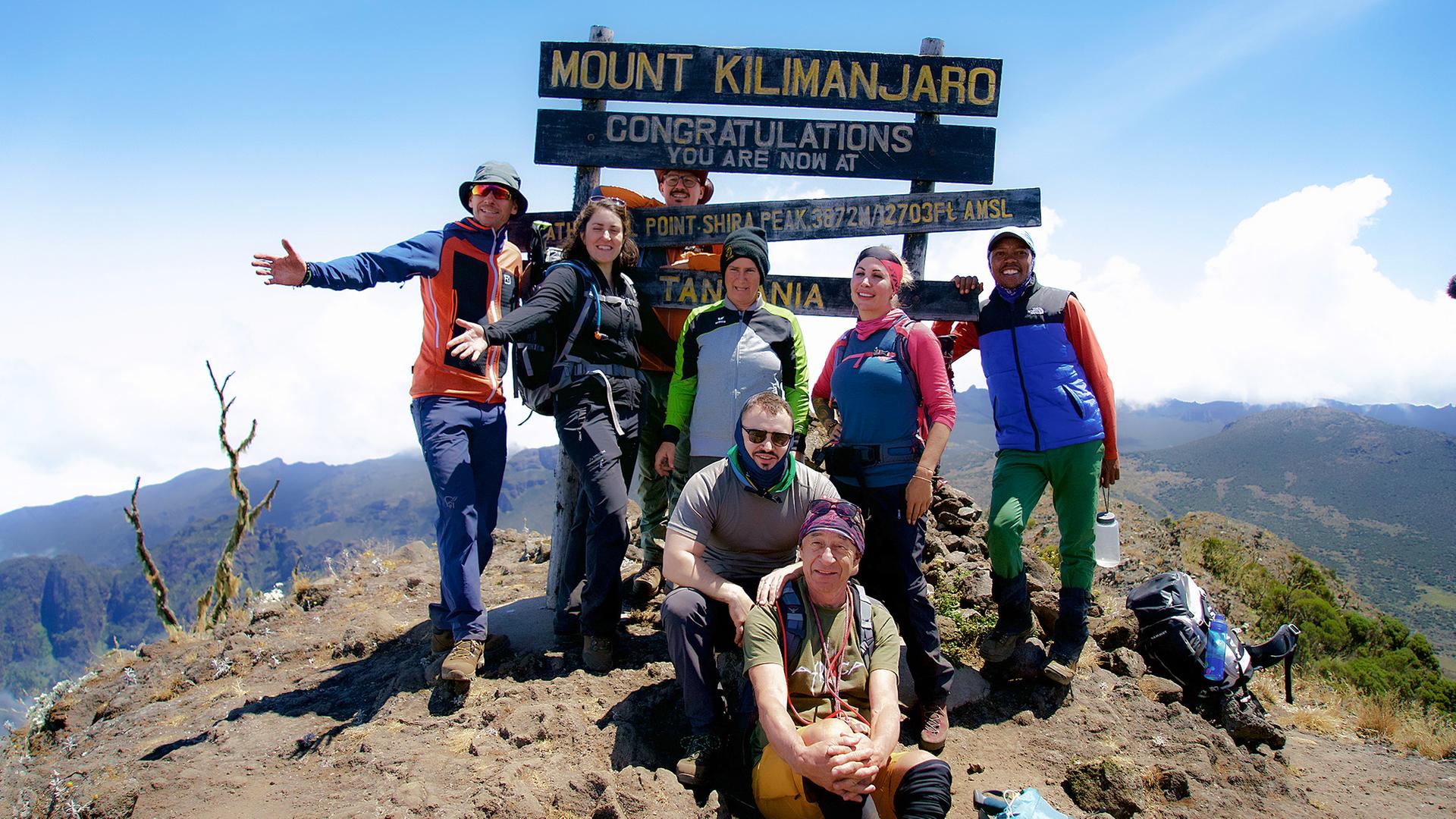 Acht Personen in Trekkingausrüstung posieren am Gipfel des Kilimandscharo für ein Gruppenfoto
