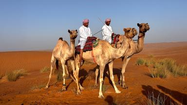 Zwei Männer in traditioneller Kleidung reiten auf Kamelen durch eine weite Wüstenlandschaft mit rotem Sand und spärlichem Grasbewuchs unter blauem Himmel.