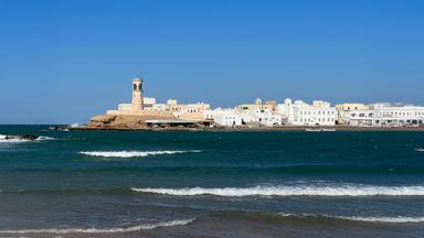 Blick über das türkisfarbene Meer auf die Küstenstadt Sur mit ihren weißen Gebäuden und dem markanten runden Leuchtturm auf einem Felsvorsprung unter wolkenlosem Himmel.