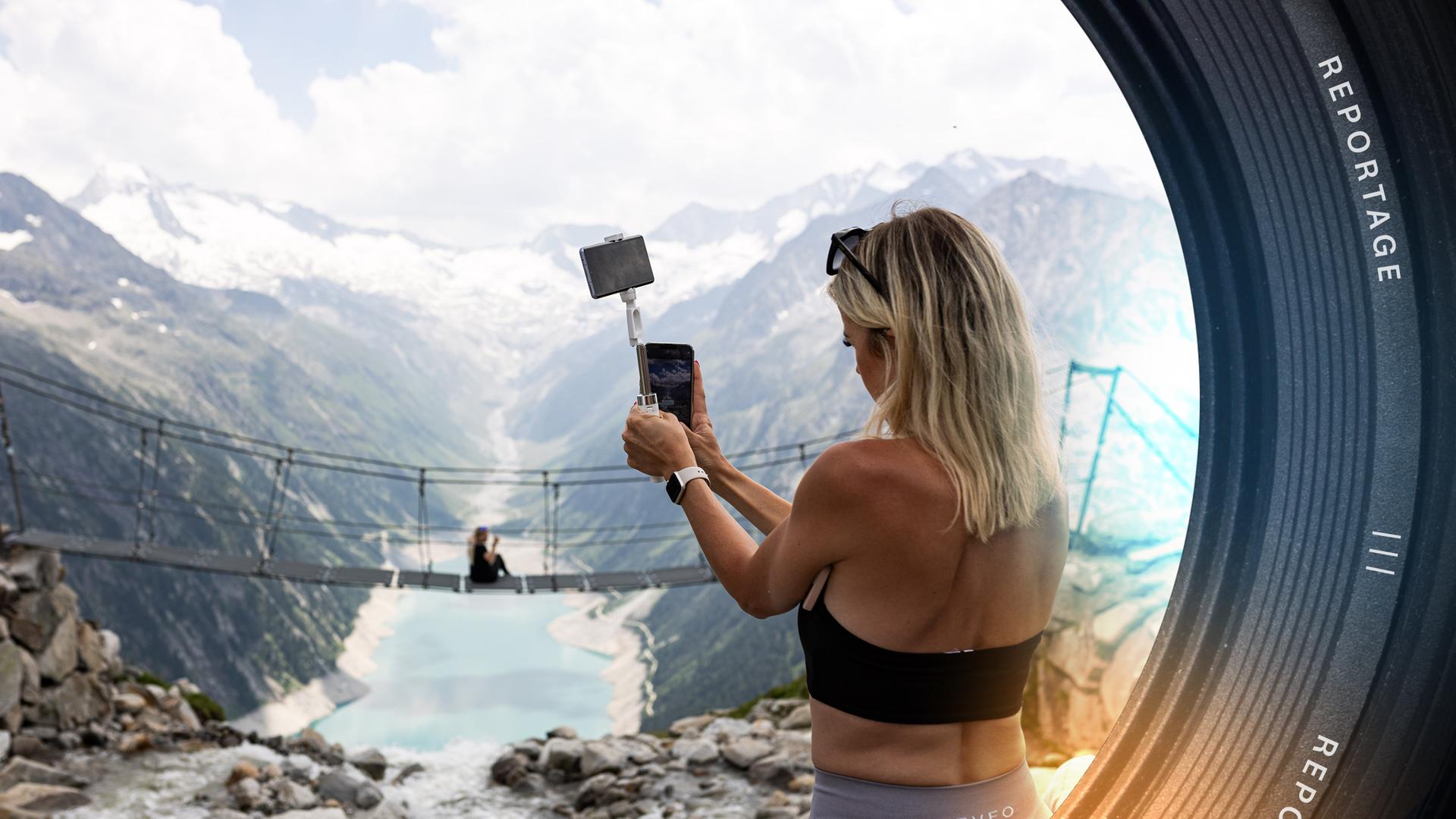 Eine junge blonde Frau steht im Vordergrund und fotografiert mit ihrem Smartphone eine Freundin auf der Hängebrücke an der Olperer Hütte vor dem Bergpanorama des Zillertals.