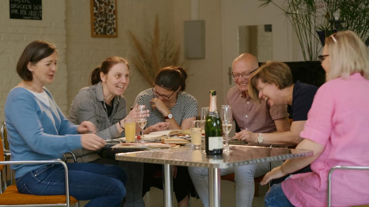 echs Erwachsene sitzen um einen Tisch herum. Es gibt Sekt, Unterlagen liegen auf dem Tisch. Sie Lachen herzlich.