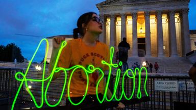 Eine Frau steht vor dem Weißen Haus und hält eine Leuchtschrift mit dem Wort "Abortion" in den Händen.