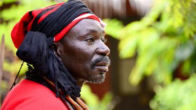 Abubakari II. in Nahaufnahme. Er trägt einen rot-schwarzen Turban und ein rotes Gewand. (Spielszene)