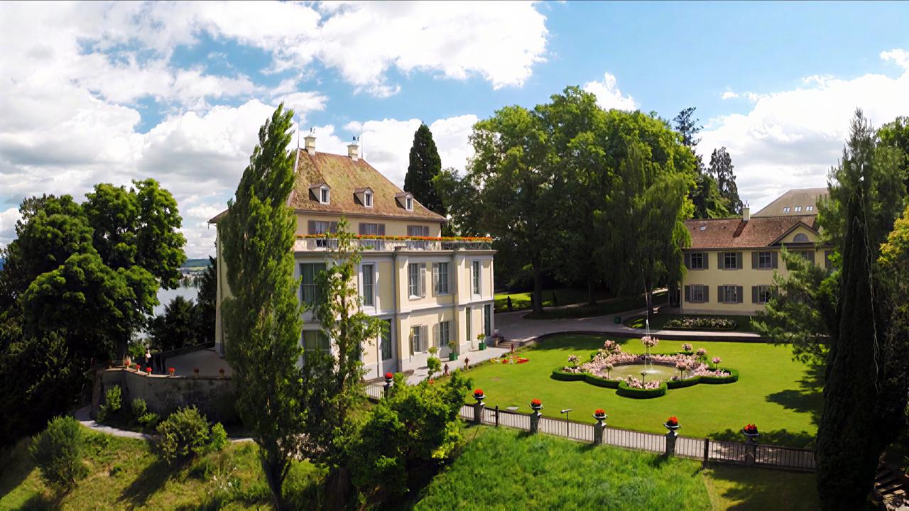 Das Bild zeigt Schloss Arenenberg in der Schweiz. Es handelt sich um ein zweigeschossiges Gebäude mit grünem Garten, umgeben von Bäumen, und einem kleineren Nebengebäude im Hintergrund.
