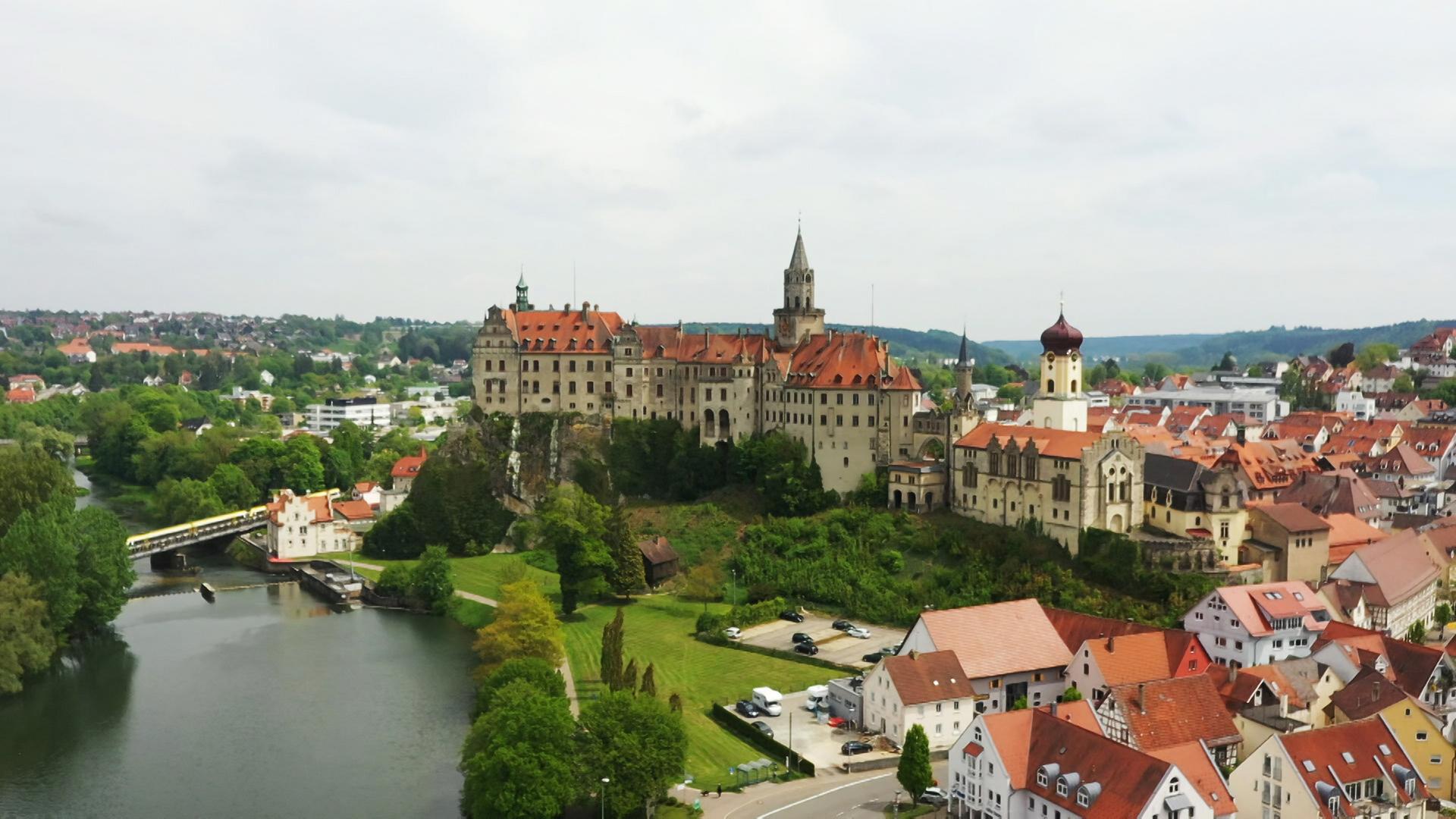Das Bild zeigt Schloss Hohenzollern in Sigmaringen, Deutschland. Das Schloss thront auf einem Hügel über einer Stadt mit roten Dächern. Umgeben ist es von Bäumen und einem Gewässer.