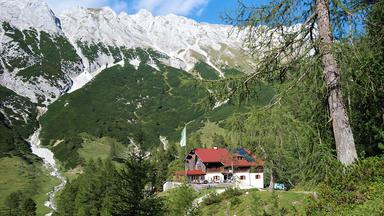 Eine Alpenvereinshütte mit rotem Dach liegt eingebettet in grüne Almwiesen und Nadelwälder vor der imposanten Kalkfelskulisse des Karwendelgebirges.